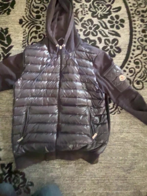 Svart dunjacka med huva från Moncler - Snygg svart dunjacka från Moncler med glansig quiltad framsida, huva och dragkedja. Ärmarna är i mjukare material och har en ficka med Moncler-logga på vänster arm. Perfekt för kyliga dagar och riktigt trendig stil. Både fickorna har håll 