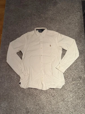 Vit skjorta från Polo Ralph Lauren - Klassisk vit skjorta från Polo Ralph Lauren med broderad logga på bröstet. Skjortan har lång ärm, knappar framtill och button-down krage. Perfekt till både jeans och chinos för en clean look.