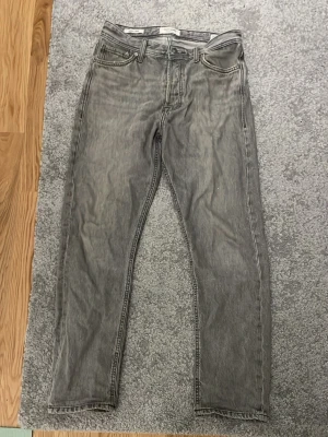 Grå jeans från Jack&Jones  - Säljer ett par grå jeans från Jack&Jones med klassisk femficksdesign och raka ben. Jeansen har en snygg tvättad look och detaljer som synliga sömmar och läderpatch bak i midjan. Perfekta för en avslappnad och trendig stil.