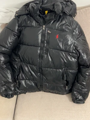 Svart dunjacka från Ralph Lauren - Snygg svart pufferjacka från Ralph Lauren med blank finish och röd logga på bröstet. Jackan har huva, dragkedja framtill och två sidofickor med dragkedja. För fler bilder och frågor skriv gärna🤝