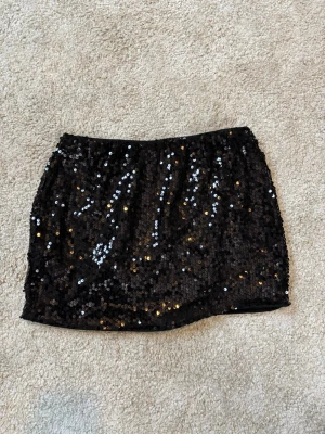 Svart paljettkjol - Snygg svart kortkjol täckt med glittriga paljetter. Perfekt för dig som vill sticka ut och glänsa lite extra. Kjolen har en rak passform och elastisk midja för skön känsla.