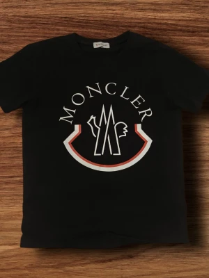 Moncler t shirt - Svart t-shirt från Moncler med stor vit och röd logga framtill. Klassisk rund halsringning och korta ärmar. Perfekt för dig som gillar stilrena och exklusiva plagg med tydlig branding.