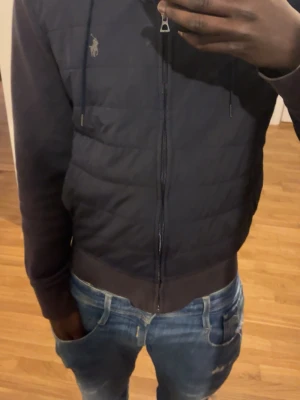 Svart cardgain från Polo Ralph Lauren - Snygg svart cardgain från Polo Ralph Lauren med dragkedja och quiltad framsida. Klassisk huva med dragsko och broderad logga på bröstet. Mjuka muddar vid ärmslut och nederkant. Perfekt för en stilren och avslappnad look.