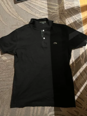 Svart pikétröja från Lacoste - Klassisk svart pikétröja från Lacoste med korta ärmar och krage. Tröjan har knäppning framtill med två knappar och den ikoniska krokodilloggan på bröstet. Tillverkad i mjuk bomull och har ett diskret mönstrat tyg på insidan av kragen.