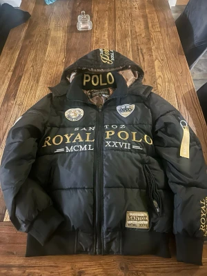 Svart pufferjacka Royal Polo SANTOZ - Svart pufferjacka från SANTOZ Royal Polo med guldfärgade broderade detaljer och stora logotexter framtill och på huvan. Jackan har huva med mönstrat innerfoder, dragkedja och flera patches. Perfekt för dig som gillar streetwear och statement-plagg.