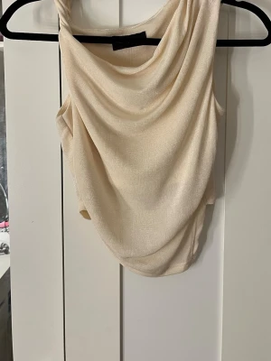 Beige draperad crop top - Trendig beige crop top med draperad framsida och asymmetrisk axel. Tillverkad i ett mjukt, stretchigt tyg som ger en snygg passform. Perfekt för att styla med höga jeans eller kjol för en modern look.