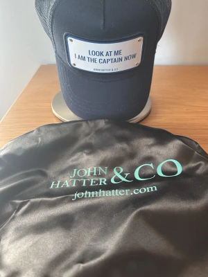 Marinblå keps från John Hatter & Co - Stilren marinblå keps med metallskylt framtill där det står 'LOOK AT ME I AM THE CAPTAIN NOW'. Justerbar baksida och klassisk böjd skärm. Perfekt accessoar för en cool och självsäker look.