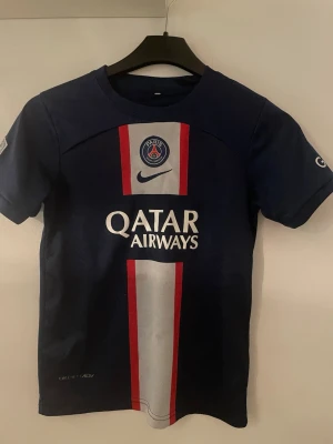 PSG Messi matchtröja blå Nike - Snygg blå PSG matchtröja från Nike med Messi och nummer 30 på ryggen. Framsidan har PSG-logga, Nike-logga och Qatar Airways-tryck samt röda och vita ränder. Perfekt för fotbollsfans som vill visa sitt stöd för laget.