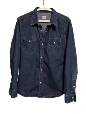 Mörkblå jeansskjorta från Lee - Klassisk mörkblå jeansskjorta från Lee med långa ärmar, tryckknappar och två bröstfickor med lock. Skjortan har en avslappnad passform och är tillverkad i slitstark bomull. Perfekt för en cool och tidlös look.