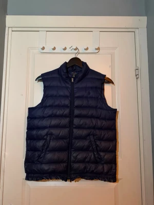 Mörkblå dunväst från Polo Ralph Lauren - Snygg mörkblå dunväst från Polo Ralph Lauren med broderad logga på bröstet. Västen har dragkedja framtill, två fickor och quiltad design. Clean look.