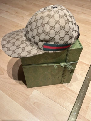 Gucci keps med GG-mönster - Säljer en beige keps från Gucci med klassiskt GG-monogram över hela kepsen och en grön-röd rand på sidan. Kepsen har böjd skärm och justerbar passform. Materialet är canvas med exklusiv känsla. Perfekt för dig som vill sticka ut med en lyxig accessoar.