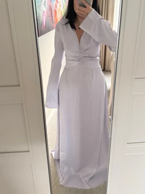 Ljuslila långklänning med v-ringning - Elegant långklänning i ljuslila med v-ringad hals och rynkdetalj framtill. Klänningen har långa, utsvängda ärmar och markerad midja för en smickrande siluett. Perfekt för dig som vill ha en stilren och feminin look.