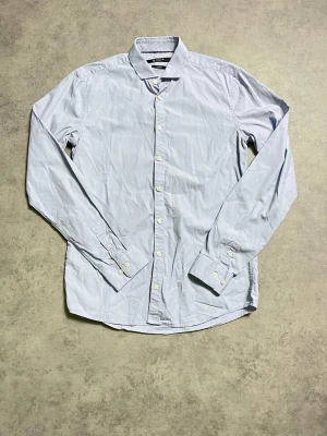 Ljusblå skjorta från Tiger of Sweden - Klassisk ljusblå skjorta från Tiger of Sweden med smal passform. Skjortan har långa ärmar, knappar framtill och är tillverkad i bomull. Perfekt för en stilren look.