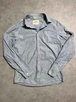 Blå skjorta från Hollister - Snygg blå långärmad skjorta från Hollister med klassisk krage och bröstficka. Skjortan har vita knappar och diskret Hollister-logga broderad på fickan. Tillverkad i ett stretchigt bomullsmaterial för extra komfort.