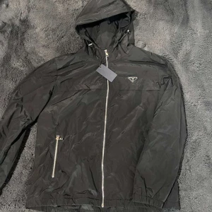 Svart vindjacka från Prada, storlek L - Svart vindjacka från Prada med huva och silvriga dragkedjor. Jackan har en clean look med Pradas ikoniska trekantslogga på bröstet. Tillverkad i lätt och glansig polyester, perfekt för dig som gillar stilren design och exklusiva märken.