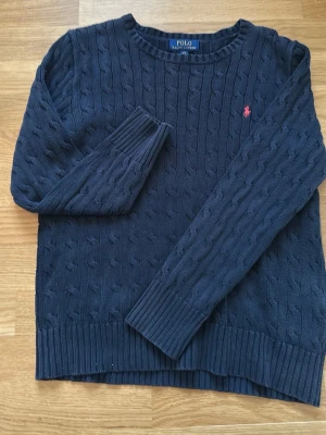 Mörkblå kabelstickad tröja Polo Ralph Lauren - Mörkblå kabelstickad tröja från Polo Ralph Lauren med rund halsringning och lång ärm. Tröjan har ribbstickade muddar och en liten röd broderad logga på bröstet. Perfekt för lager-på-lager och klassisk stil.
