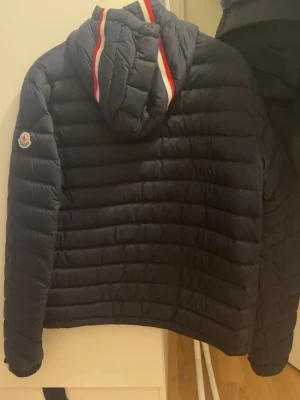 Mörkblå dunjacka från Moncler - Snygg mörkblå dunjacka från Moncler med huva och dragkedja framtill. Jackan har quiltad design, två fickor med dragkedja och en ikonisk Moncler-logga på ärmen. Huva med röd och vit randig detalj. Original pris ligger på 11300. 