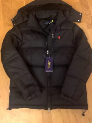 Svart dunjacka från Polo Ralph Lauren - Svart dunjacka från Polo Ralph Lauren med röd broderad logga på bröstet. Jackan har dragkedja framtill, huva med tryckknappar och klassisk puffer-design. Perfekt för kalla dagar och riktigt snygg streetstil. Helt ny 