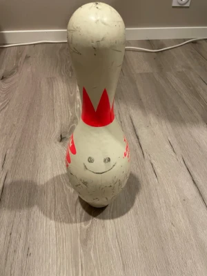 Bowlingkägla från Brunswick - En klassisk vit bowlingkägla från Brunswick med röd dekor och tryck. Käglan har en typisk avlång form och är tillverkad i hård plast. På framsidan finns ett handritat leende ansikte. Perfekt som dekoration eller samlarobjekt.
