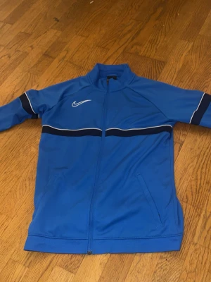 Blå träningsjacka från Nike XL BARNSTORLEK - Snygg blå träningsjacka från Nike med dragkedja framtill, vita och svarta detaljer över bröstet och ärmarna samt två fickor. Jackan har hög krage och är tillverkad i ett mjukt, sportigt material som passar perfekt till träning eller uppvärmning.