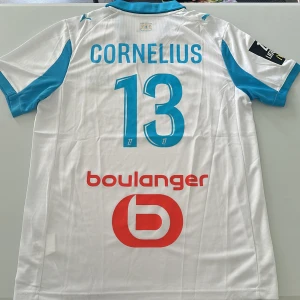 Marseille Cornelius matchtröja Puma - Säljer en Olympique de Marseille fotbollströja från Puma med Cornelius och nummer 13 på ryggen. Tröjan är vit med ljusblå detaljer, broderat klubbmärke och sponsortryck. Tillverkad i ventilerande polyester med korta ärmar och klassisk krage. Replika, endast handtvätt gäller!