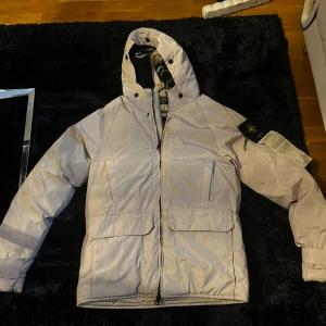Ljusbeige dunjacka från Stone Island S - Säljer en ljusbeige dunjacka från Stone Island med huva och klassisk logotyp på ärmen. Jackan är quiltad, har stora fickor framtill och dragkedja. passar perfekt för kyliga dagar. Jackan passar även som en liten M