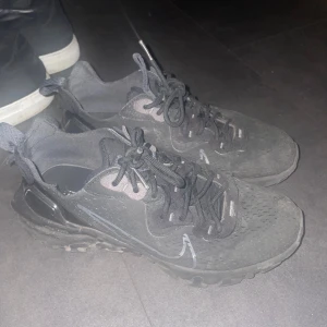 Svarta Nike React sneakers - Nike React sneakers i svart med grå detaljer och reflekterande swoosh på sidan. Skorna har en chunky, mönstrad sula och ventilerande mesh-ovandel. Klassisk snörning och låg profil. Perfekt för dig som gillar sportig och modern stil. Se bild 3