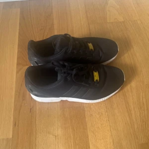 Svarta Adidas sneakers med Torsion-sula - Svarta Adidas sneakers med klassiska tre ränder på sidan och vit sula. Skorna har snörning och detaljer i gult på tungan och under sulan där det står Torsion. Ovandelen är i mesh-liknande syntetmaterial för bra andningsförmåga.