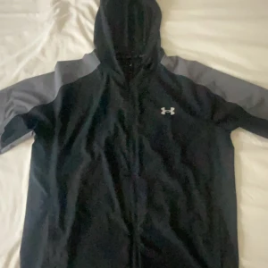 Svart och grå vindjacka Under Armour - Under Armour vindjacka i svart med grå kontrast på ärmarna och huva. Jackan har dragkedja framtill och en snygg logga på bröstet. Perfekt för träning eller chill dagar. Materialet är lätt och syntetiskt, designad för att vara bekväm och funktionell.