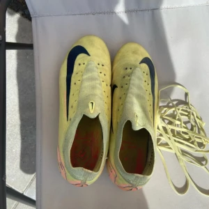 Gula Nike fotbollsskor med svart swoosh - Säljer ett par gula Nike fotbollsskor med svart swoosh och orange detaljer. Skorna har en strömlinjeformad design, platt sula med dobbar och är tillverkade i syntetmaterial. Skosnören i matchande gul färg medföljer. Perfekta för dig som vill sticka ut på planen.