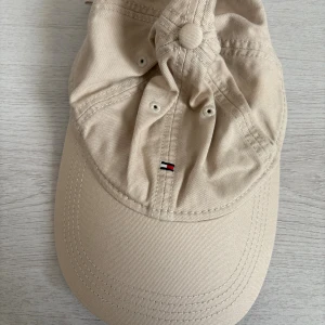 Beige keps från Tommy Hilfiger - Snygg beige keps från Tommy Hilfiger med klassisk böjd skärm och broderad logga framtill. Kepsen är tillverkad i bomull och har ventilerande hål samt justerbar passform. Perfekt accessoar för en clean och stilren look.