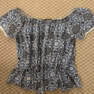 Blå offshoulder blus med mönster - Säljer en blå och vit offshoulder blus med korta ärmar och ett detaljerat blommigt mönster. Blusen har en elastisk halsringning och små dekorativa bollar längs kanten. Perfekt för dig som gillar boho-vibe och snygga prints.