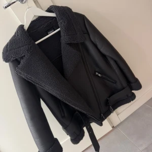 Svart jacka från ONLY med bälte - Säljer en svart  jacka från ONLY med stora kragar, mysigt teddyfoder och detaljer som bälte i midjan och dragkedjor. Endast använt den fåtal gånger.