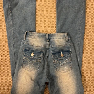 Blå jeans med dekorerade bakfickor - Snygga blå jeans med slitningar och dekorerade bakfickor med silverfärgade knappar och broderade detaljer. Jeansen har normal passform och klassisk femficksdesign. Perfekta för dig som vill ha ett par jeans med lite extra detaljer.