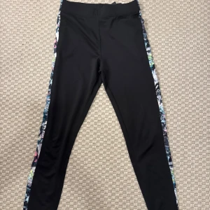 Svarta leggings med färgglada sidor - Svarta leggings med färgglada, abstrakta mönster längs sidorna. Tight passform och hög midja, perfekta för träning eller chill. Materialet är stretchigt och mjukt, troligtvis polyester eller syntet. Snygg detalj med de färgsprakande panelerna som ger extra edge.