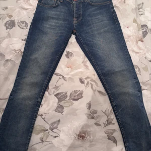 Blå jeans från Nudie Jeans Co, W30 L30 - Säljer ett par klassiska blå jeans från Nudie Jeans Co i modellen W30 L30. Jeansen har orangea sömmar, fem fickor och snygg slitning framtill. Materialet är jeans och passformen är rak med normal midja. Perfekta för en avslappnad och stilren look.