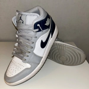 Nike Air Jordan 1 Mid vit/grå/blå - Nike Air Jordan 1 Mid sneakers i storlek 44 med snygg färgkombo av vitt, grått och marinblått. Klassisk Jordan-logga på sidan och tungan, perforerad tå och snörning. Skorna har en rund tå och platt sula, tillverkade i skinn och syntet för en clean och sportig look.