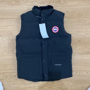 Svart dunväst från Canada Goose (helt ny+gratis frakt) - Svart dunväst från Canada Goose med klassisk logga på bröstet och två stora fickor framtill. Västen har hög krage och knäppning med svarta knappar. Perfekt för kalla dagar och har en clean, stilren look.