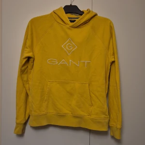 Gul hoodie från GANT med broderad logga - Gul hoodie från GANT med broderad vit logga på bröstet. Tröjan har huva, känguruficka och långa ärmar. Ett litet hål på fickan, annars i fint använt skick. 