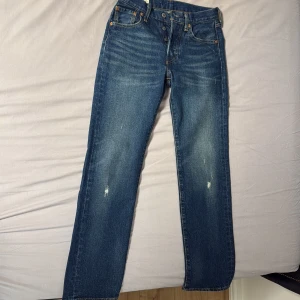 Levi's 501 blå jeans W26 L30 - Klassiska Levi's 501 jeans i blå denim med raka ben och knäppning med knappar. Snyggt slitna detaljer framtill och bak, samt ikonisk läderpatch och röd Levi's-tagg på bakfickan. Perfekta för en avslappnad och tidlös look.Som helt nya nästan aldrig använda då dem är små 