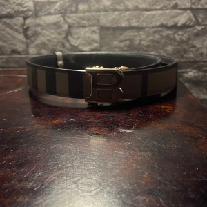 Randigt Burberry bälte med guldspänne - Snyggt bälte från Burberry med breda svarta och beige ränder och ett stort B-format spänne i guld. Bältet är gjort i skinn och har en klassisk, stilren design som sticker ut med sitt ikoniska mönster och exklusiva känsla.
