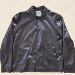 Svart skinnjacka från Zara - Stilren svart skinnjacka från Zara med klassisk krage och raka linjer. Jackan har knäppning framtill, två sidofickor och en clean look som passar till allt. Materialet är glansigt och ger en modern vibe.