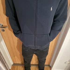 Mörkblå hoodie från Polo Ralph Lauren - Säljer en mörkblå hoodie från Polo Ralph Lauren med dragkedja och klassisk logga på bröstet. Tröjan har huva med snörning, känguruficka och långa ärmar. Perfekt för en chill och stilren look.