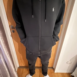 Svart hoodie från Polo Ralph Lauren - Svart hoodie från Polo Ralph Lauren med dragkedja och huva. Klassisk design med vit broderad logga på bröstet, kängurufickor och snörning i huvan. Tillverkad i mjuk bomull för en skön och avslappnad känsla.