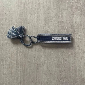 Blå nyckelring med tofsar - Säljer en blå och vit nyckelring i tyg med texten 'CHRISTIAN DIOR J'ADIOR'. Nyckelringen har flätad ögla och två dekorativa tofsar. Perfekt som accessoar till väskan eller nycklarna.