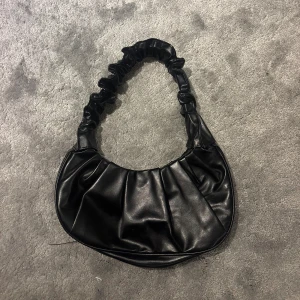 Svart axelväska i skinnimitation - Trendig svart axelväska med rynkad rem och rundad form. Väskan är gjord i glansig skinnimitation och har magnetlås. Perfekt storlek för det viktigaste och ger en cool vibe till din outfit.