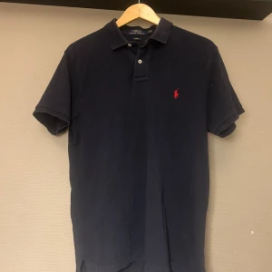 Ralph Lauren pikétröja  - Snygg marinblå pikétröja från Polo Ralph Lauren i slim fit-modell. Klassisk krage, korta ärmar och två knappar framtill. Ikoniska röda loggan broderad på bröstet. Tillverkad i mjuk bomull som är skön mot huden. Ny pris 1200