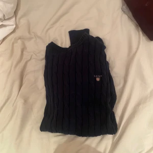 Mörkblå kabelstickad tröja från Gant - Kabelstickad mörkblå tröja från Gant i storlek S. Tröjan har rund halsringning, ribbade muddar och Gant-logga broderad på bröstet. Klassisk och stilren design som funkar till det mesta.