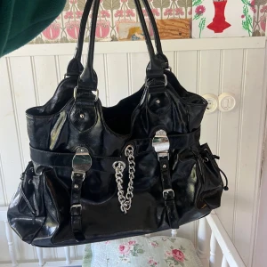Svart axelväska med kedjedetaljer - Säljer en snygg svart axelväska i skinnliknande material. Väskan har coola kedjedetaljer och spännen på framsidan, vilket ger den en edgy look. Perfekt för både vardag och fest! Den har också praktiska dragkedjor och rymliga fack för alla dina grejer