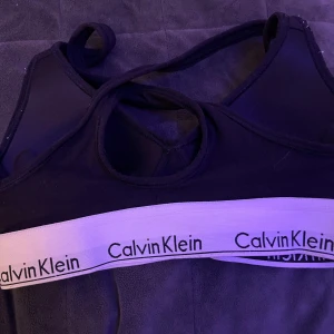 Svart Calvin Klein linnetopp - Svart bh från Calvin Klein med korsade axelband och bred vit resår med logotypen tryckt runt om. Snygg och sportig design som ger en cool vibe. Materialet är mjuk bomull med lite stretch för skön passform.🖤🖤